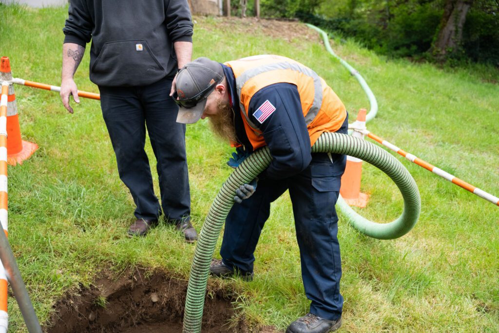 The image shows a man in uniform working on a septic tank, handling a large hose with care and precision. His focused expression and the specialized equipment suggest that he is performing a crucial maintenance or pumping task. The scene highlights the hands-on, expert service provided by the company, emphasizing their capability to manage essential septic system needs. This image is particularly relevant to homeowners in Washington State, especially in King and Pierce County, who require reliable septic services to ensure the proper functioning of their systems. The visual underscores the company’s commitment to thorough, professional service, ensuring that septic tanks are properly maintained to prevent issues. It reflects the importance of expertise and the use of appropriate tools in delivering high-quality septic solutions to the community.